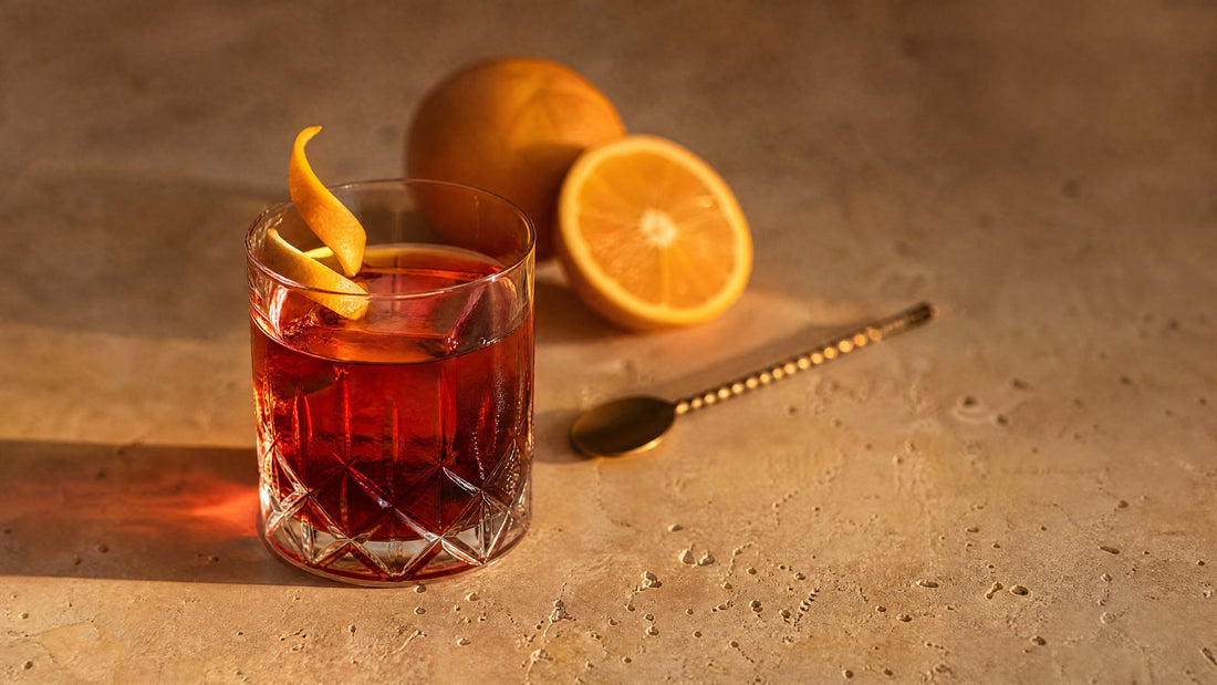 Negroni mit großem Eiswürfel und Orangenzeste im Tumbler auf Travertin-Tisch