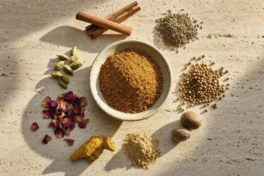 Ras el Hanout Gewürzmischung in einer Schale, daneben Zimt, Kreuzkümmel, Koriander, Kardamom, Rosenblüten, Kurkuma, Ingwer und Muskatnuss